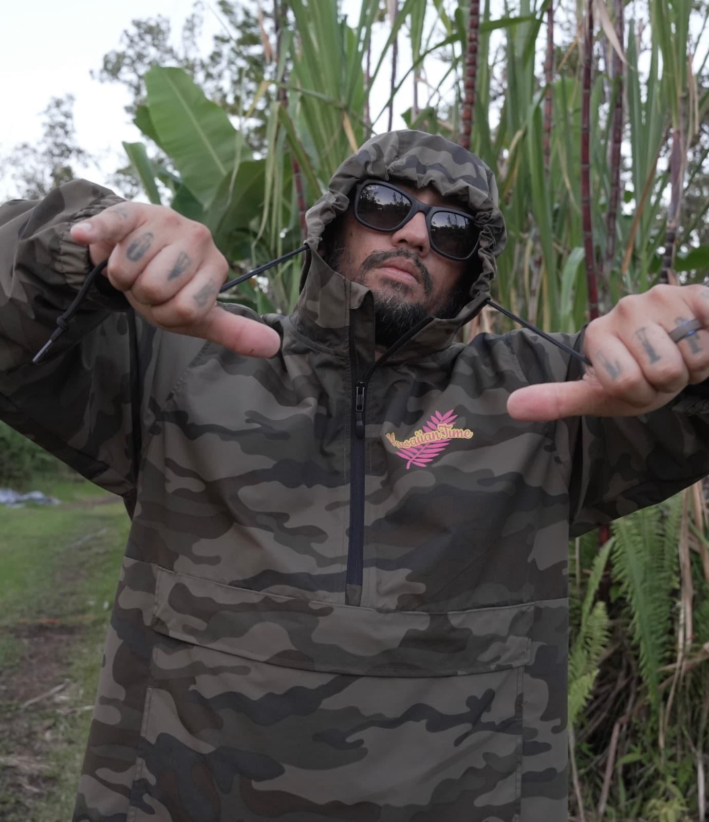 John in a hooded camo anorak and dark sunglasses, both thumbs pointing in at himself, with tall sugar cane and banana leaves behind him.